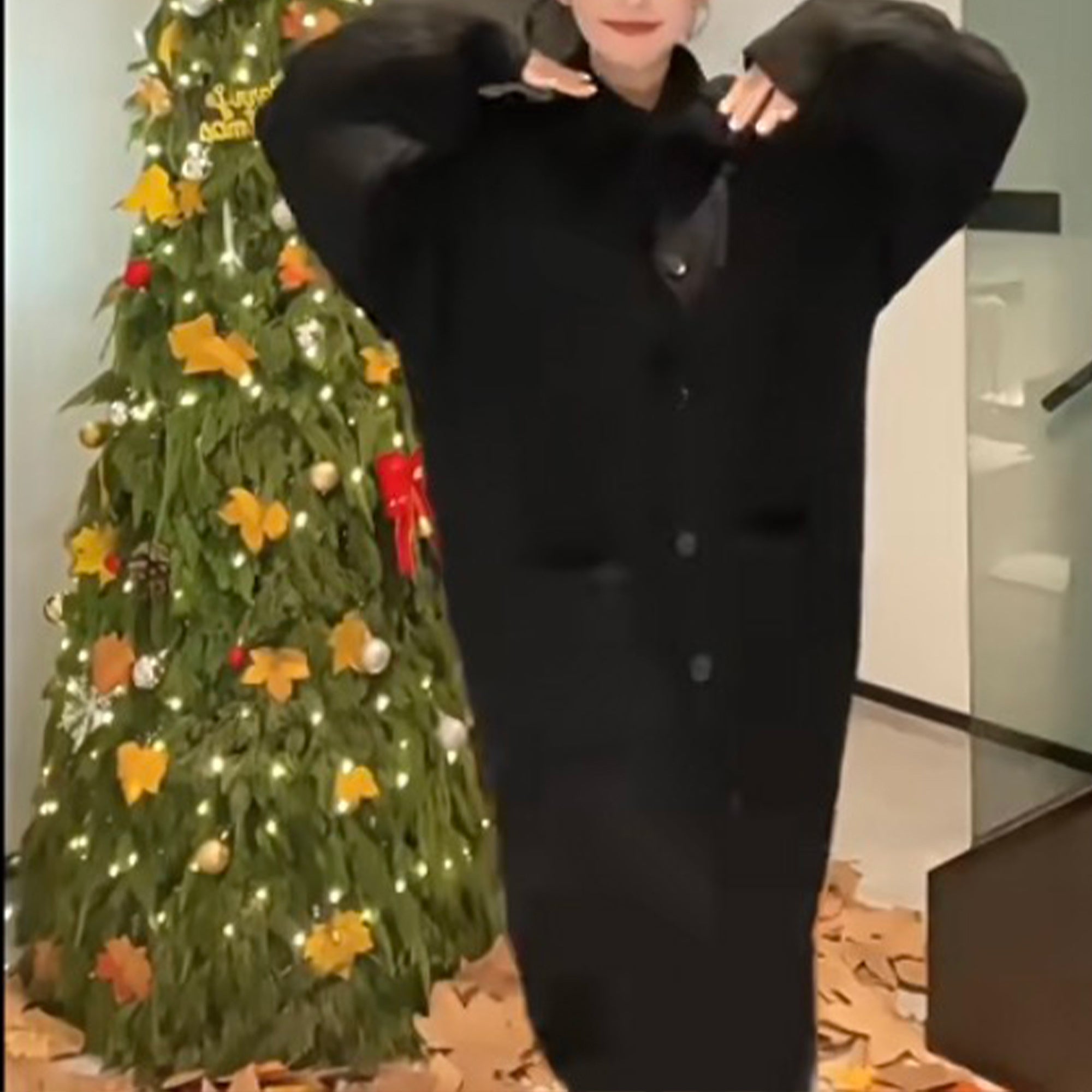 Woman wearing a cozy black cardigan in front of a decorated Christmas tree with lights.